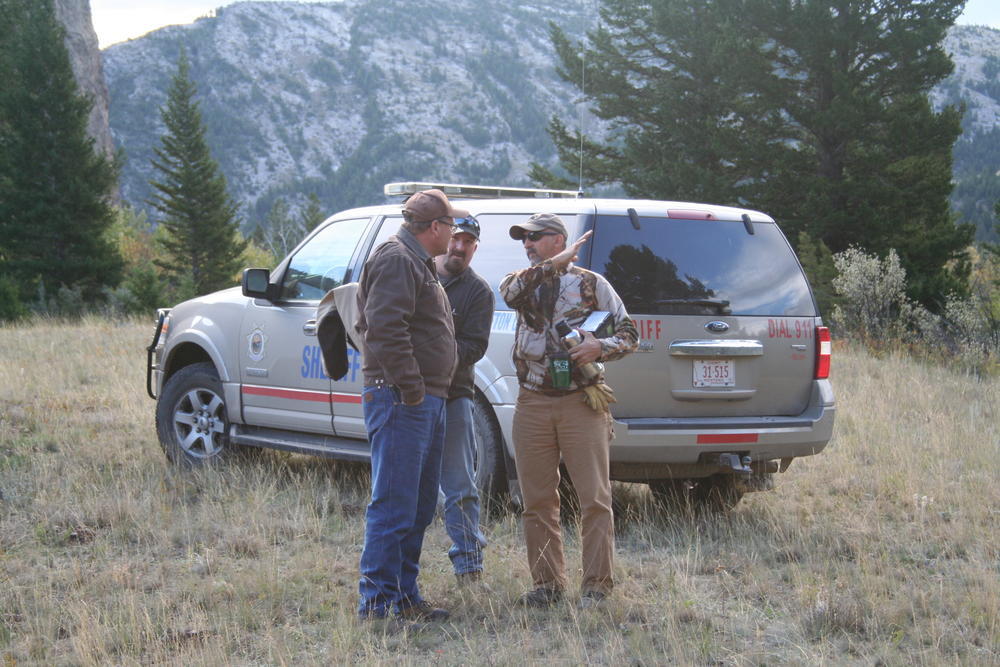 3 men talking by a sheriff's vehicle, one using his arm to indicate the angle of a slope