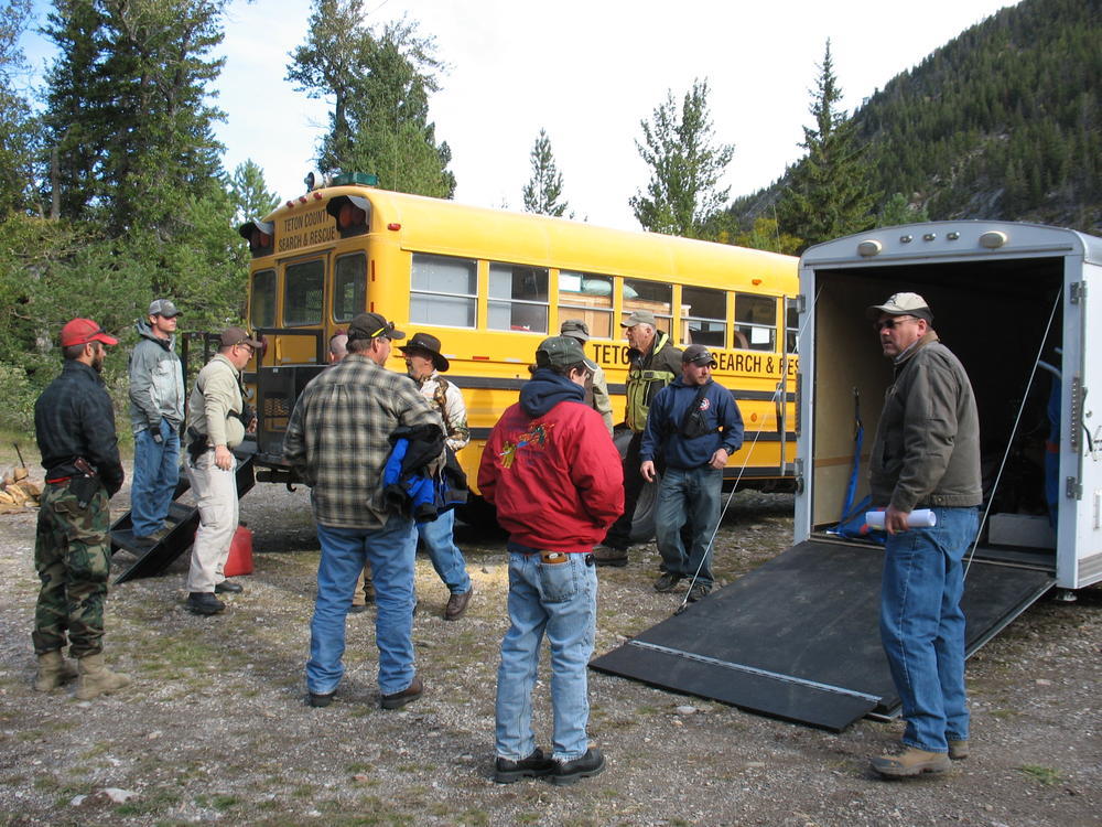 several people standing outside a school bus and a trailer with the tailgate down